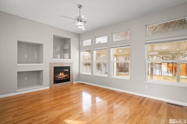 unfurnished living room with a tiled fireplace, built in shelves, ceiling fan, and light wood-type flooring