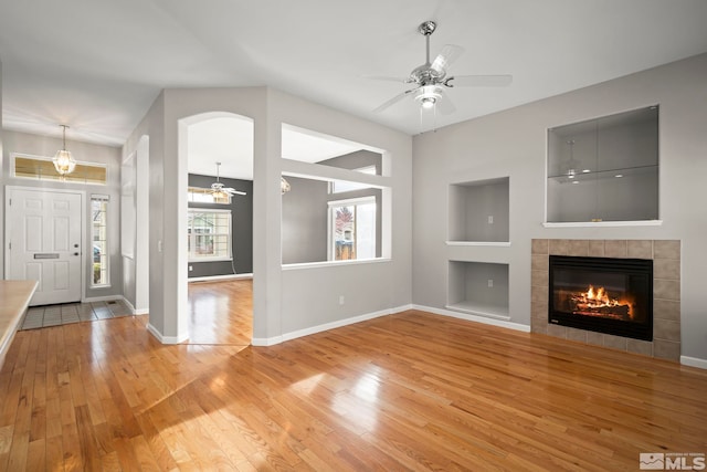 unfurnished living room with a tiled fireplace, built in shelves, ceiling fan, and light wood-type flooring