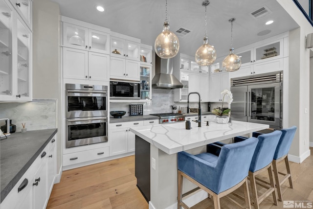 kitchen with dark stone countertops, white cabinets, built in appliances, a center island with sink, and wall chimney exhaust hood