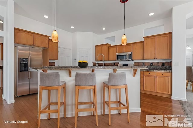 kitchen with wood-type flooring, decorative light fixtures, dark stone countertops, an island with sink, and stainless steel appliances