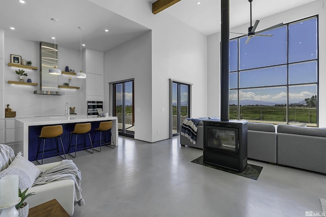 living room featuring ceiling fan, beam ceiling, concrete flooring, a high ceiling, and a wood stove