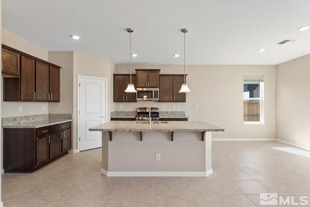 kitchen with sink, a kitchen island with sink, hanging light fixtures, dark brown cabinets, and light stone counters