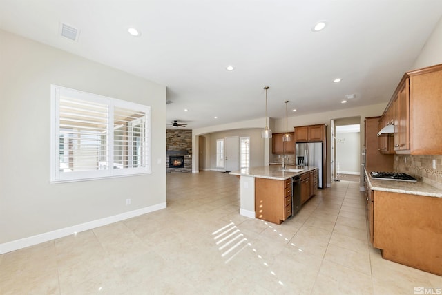 kitchen featuring a stone fireplace, pendant lighting, an island with sink, backsplash, and light stone countertops