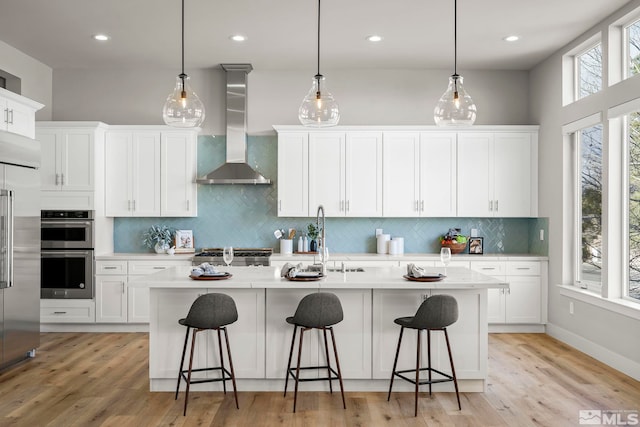 kitchen with wall chimney exhaust hood, hanging light fixtures, a center island with sink, and white cabinets