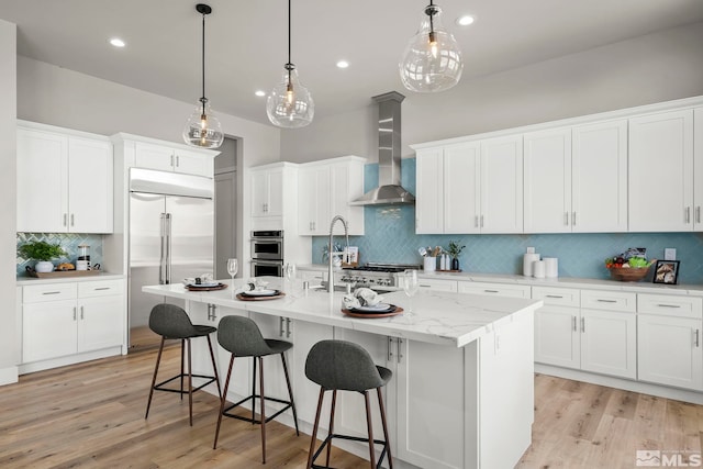 kitchen featuring white cabinetry, appliances with stainless steel finishes, wall chimney exhaust hood, and an island with sink