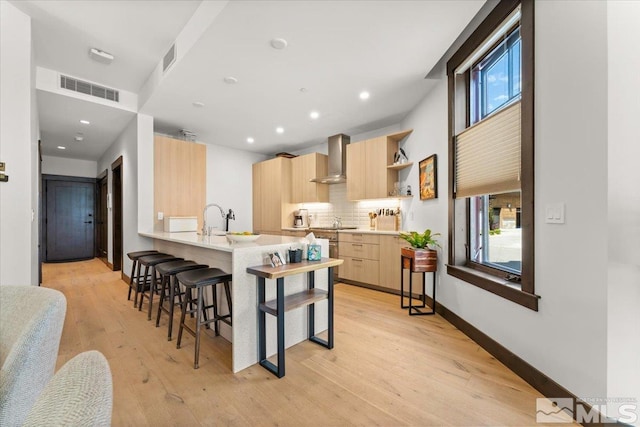 kitchen with decorative backsplash, kitchen peninsula, light brown cabinets, light wood-type flooring, and wall chimney exhaust hood