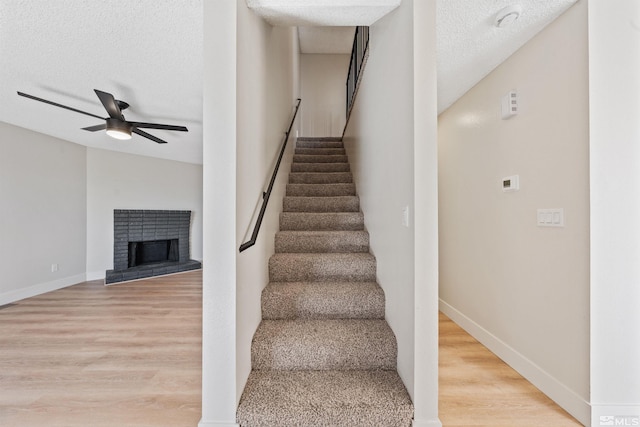 staircase with hardwood / wood-style flooring, ceiling fan, a brick fireplace, and a textured ceiling