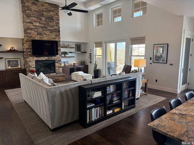 living room with a high ceiling, ceiling fan, a stone fireplace, and dark hardwood / wood-style flooring