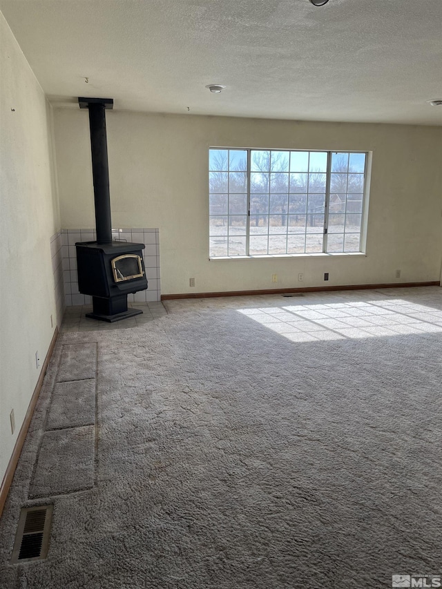 unfurnished living room with a textured ceiling, light colored carpet, visible vents, baseboards, and a wood stove