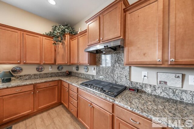 kitchen featuring under cabinet range hood, light stone counters, brown cabinets, and stainless steel gas cooktop