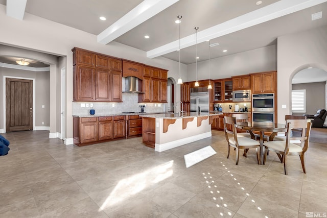 kitchen featuring built in appliances, wall chimney exhaust hood, an island with sink, and arched walkways