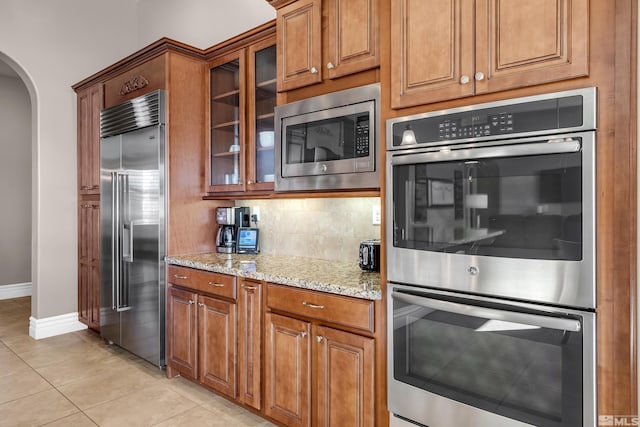 kitchen with built in appliances, arched walkways, light stone counters, brown cabinets, and glass insert cabinets