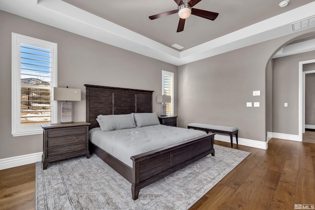 bedroom with arched walkways, a raised ceiling, baseboards, and wood finished floors