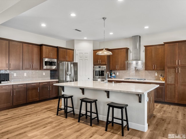 kitchen featuring an island with sink, wall chimney exhaust hood, a kitchen breakfast bar, light stone countertops, and stainless steel appliances