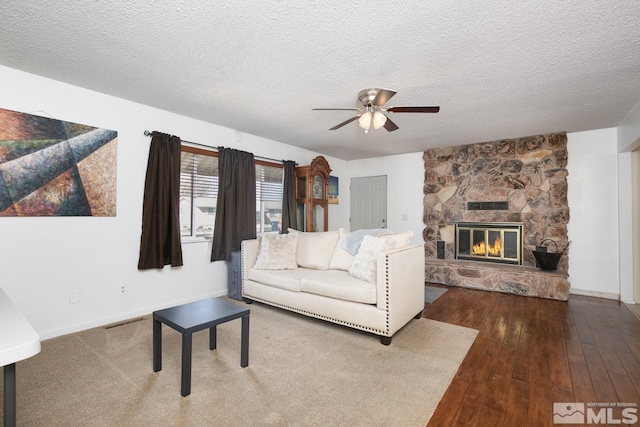 living room with a ceiling fan, a stone fireplace, a textured ceiling, and wood finished floors