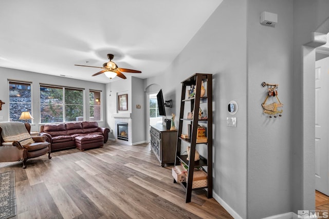 living room featuring ceiling fan, a glass covered fireplace, light wood-style flooring, and baseboards