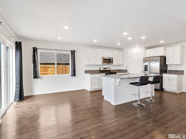 kitchen featuring stainless steel appliances, white cabinets, a kitchen island with sink, dark stone counters, and a kitchen breakfast bar