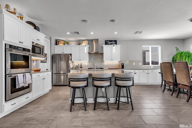 kitchen featuring a center island with sink, visible vents, appliances with stainless steel finishes, hanging light fixtures, and wall chimney range hood