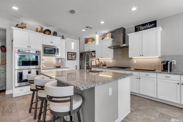 kitchen featuring visible vents, an island with sink, wall chimney exhaust hood, appliances with stainless steel finishes, and decorative light fixtures