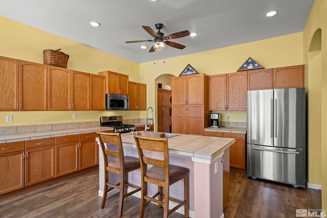 kitchen featuring arched walkways, a breakfast bar area, a kitchen island with sink, stainless steel appliances, and light countertops