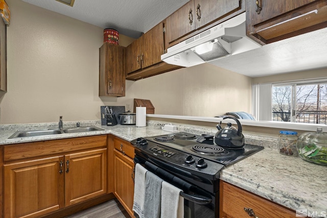 kitchen with a textured ceiling, under cabinet range hood, black range with electric stovetop, a sink, and brown cabinetry