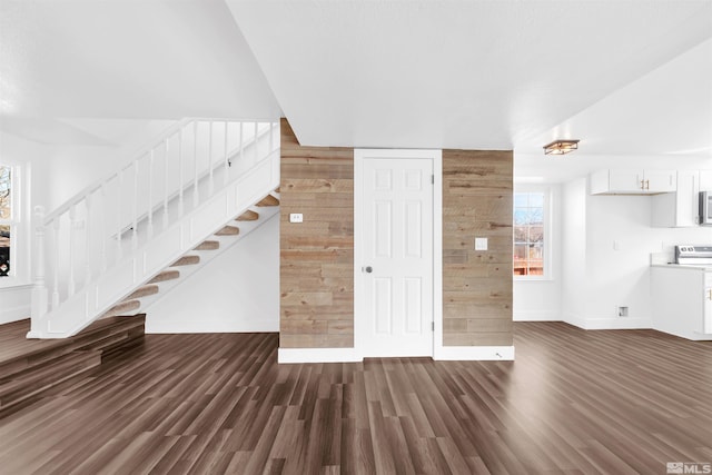 unfurnished living room with stairs, wooden walls, baseboards, and dark wood-style flooring