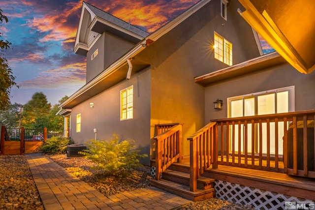 property exterior at dusk featuring central AC, fence, a deck, and stucco siding