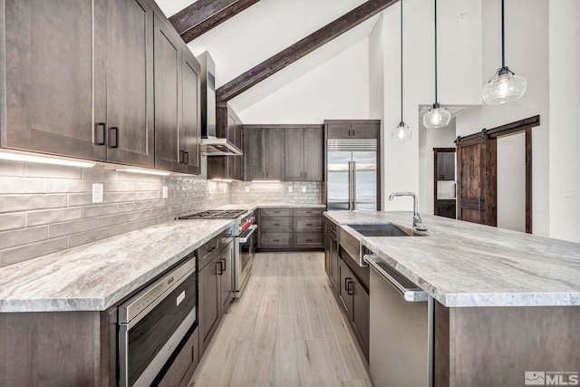 kitchen with built in appliances, a barn door, a sink, wall chimney exhaust hood, and decorative light fixtures