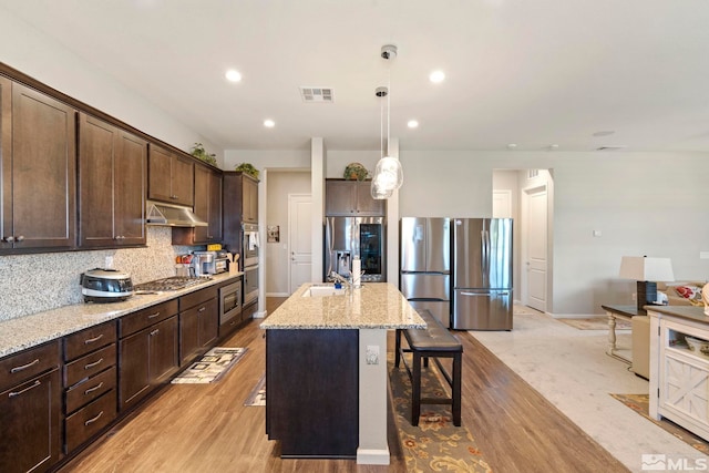 kitchen featuring light stone counters, a breakfast bar, a center island with sink, stainless steel appliances, and under cabinet range hood