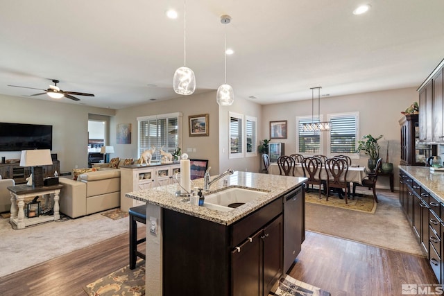 kitchen featuring decorative light fixtures, a kitchen island with sink, a sink, dark brown cabinets, and light stone countertops