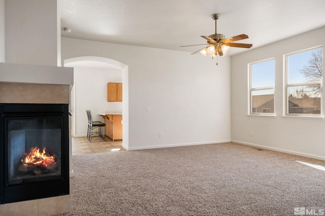 unfurnished living room with arched walkways, a fireplace, light colored carpet, a ceiling fan, and baseboards