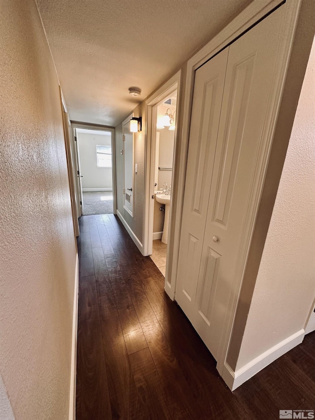 hallway featuring a textured ceiling, a textured wall, dark wood finished floors, and baseboards