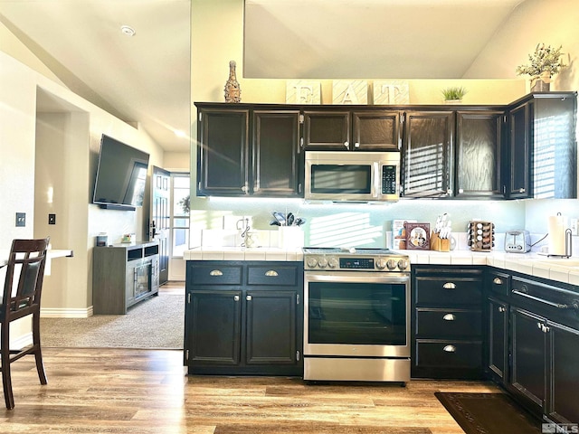 kitchen featuring light wood-type flooring, tile countertops, stainless steel appliances, baseboards, and vaulted ceiling