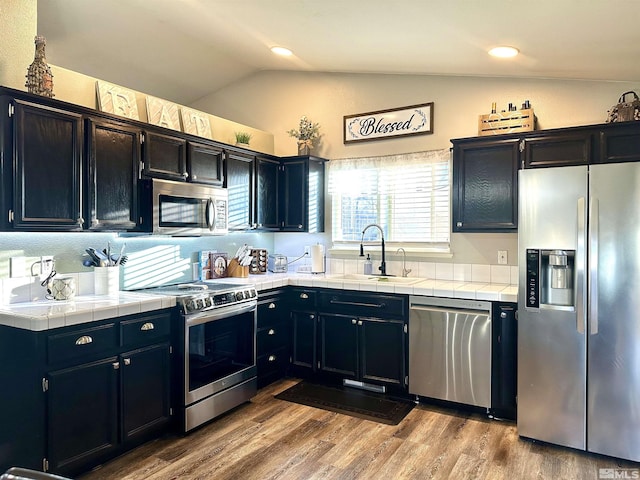 kitchen with a sink, wood finished floors, appliances with stainless steel finishes, tile counters, and vaulted ceiling