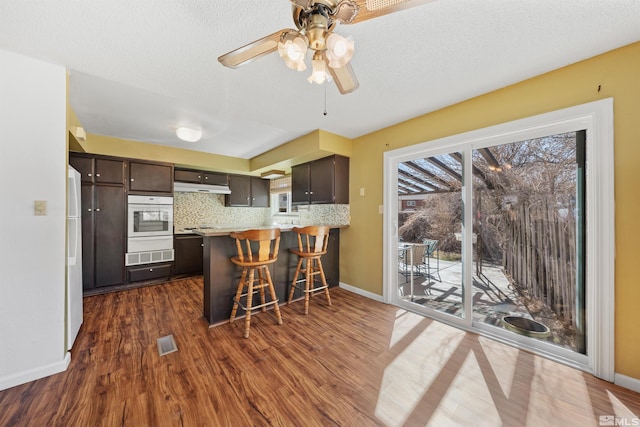 kitchen featuring white appliances, dark wood finished floors, a peninsula, dark brown cabinets, and backsplash