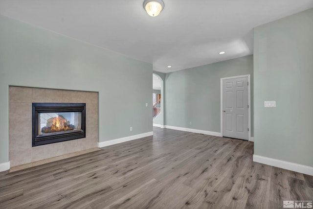 unfurnished living room with a tile fireplace, arched walkways, baseboards, and wood finished floors