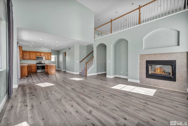 unfurnished living room featuring light wood-style flooring, a high ceiling, a tiled fireplace, baseboards, and stairs