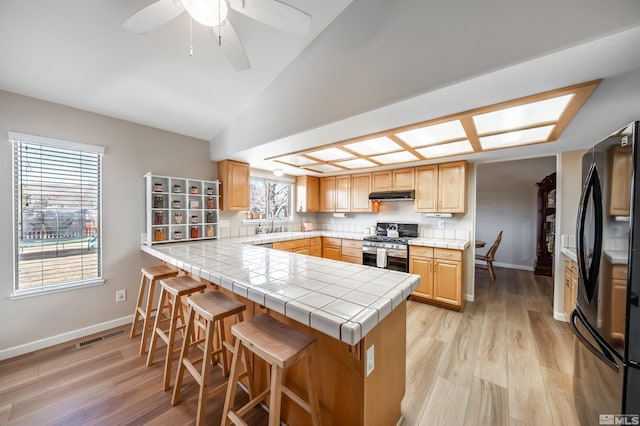 kitchen with a breakfast bar, freestanding refrigerator, a peninsula, under cabinet range hood, and stainless steel gas range oven