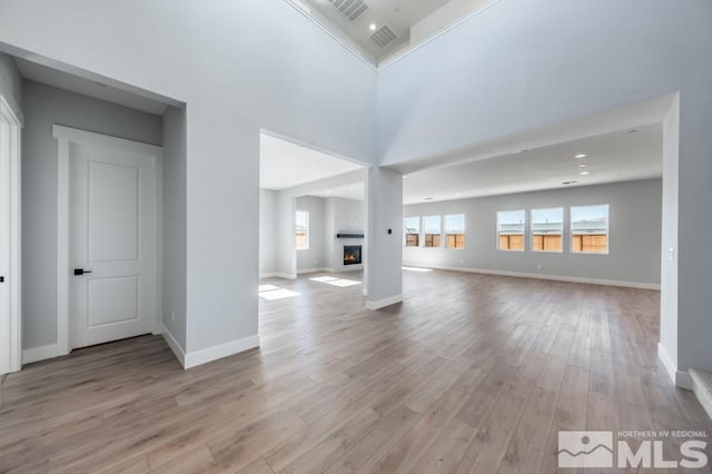 unfurnished living room featuring a warm lit fireplace, a towering ceiling, visible vents, baseboards, and light wood-type flooring