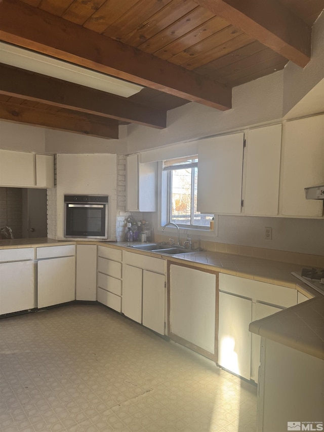 kitchen with beam ceiling, tile countertops, light floors, stainless steel oven, and a sink