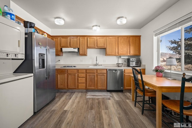 kitchen featuring under cabinet range hood, stainless steel appliances, stacked washer and dryer, wood finished floors, and a sink