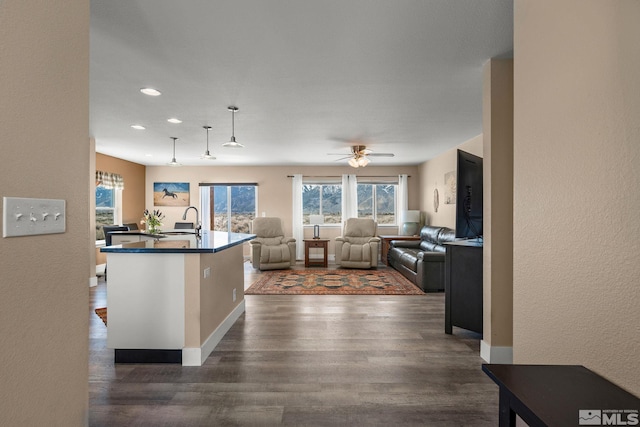 kitchen featuring dark wood finished floors, dark countertops, ceiling fan, a kitchen island with sink, and a sink