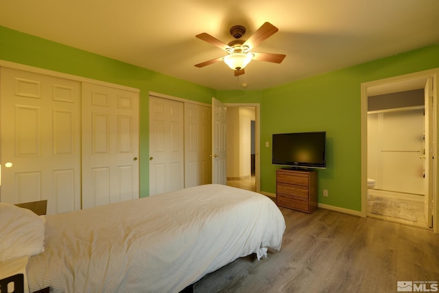 bedroom featuring baseboards, a ceiling fan, ensuite bath, wood finished floors, and two closets