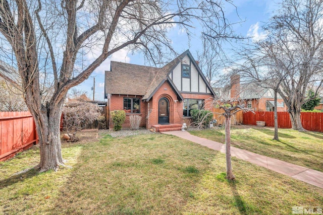 tudor-style house with a shingled roof, a front yard, fence, and brick siding