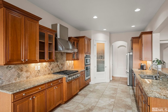 kitchen featuring arched walkways, stainless steel appliances, a sink, light stone countertops, and wall chimney exhaust hood