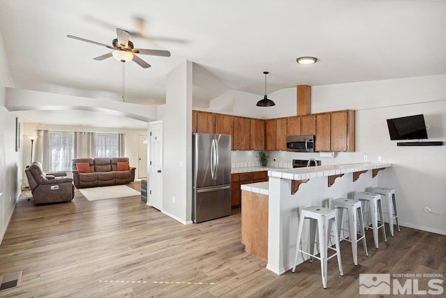 kitchen featuring brown cabinetry, tile countertops, appliances with stainless steel finishes, a breakfast bar area, and a peninsula