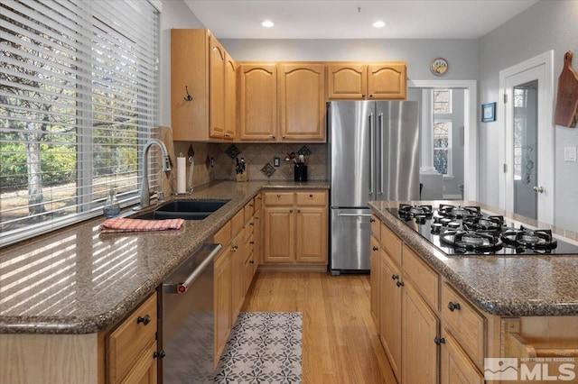 kitchen with stainless steel appliances, a sink, light wood-style floors, backsplash, and dark stone counters