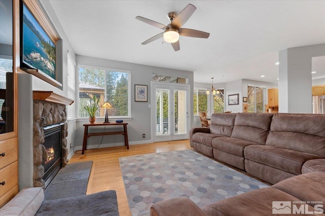 living area featuring light wood-style flooring, a stone fireplace, baseboards, and ceiling fan with notable chandelier