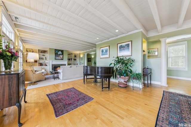 interior space with a warm lit fireplace, baseboards, beam ceiling, light wood finished floors, and decorative columns