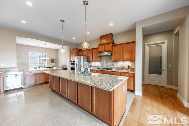 kitchen with a center island with sink, brown cabinetry, appliances with stainless steel finishes, light stone countertops, and under cabinet range hood